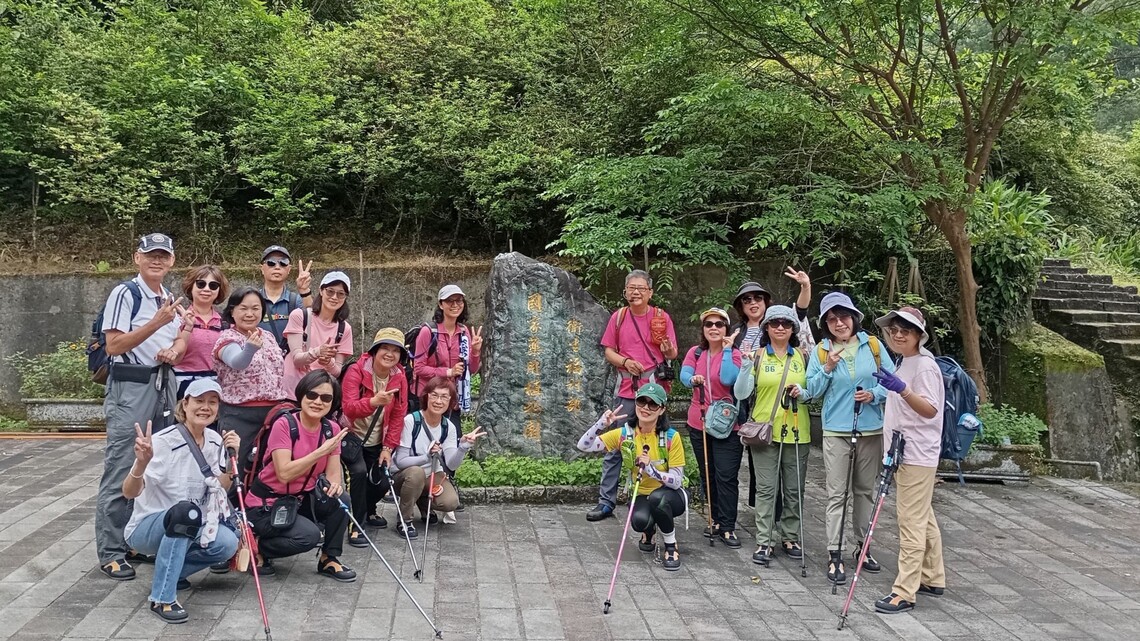 照片攝於 2025 年 5 月 2 日，植物共學班的 20 位成員於「衛生福利部 國家藥用植物園」石碑前合影留念。參訪成員多為女性，穿著休閒或登山裝備，配戴帽子與背包，部分人持登山杖。大部分人面帶笑容、擺出比 V 手勢，展現活力與團隊情誼。背景為綠意盎然的樹林與階梯，地面為灰色磚鋪地，天氣涼爽宜人。