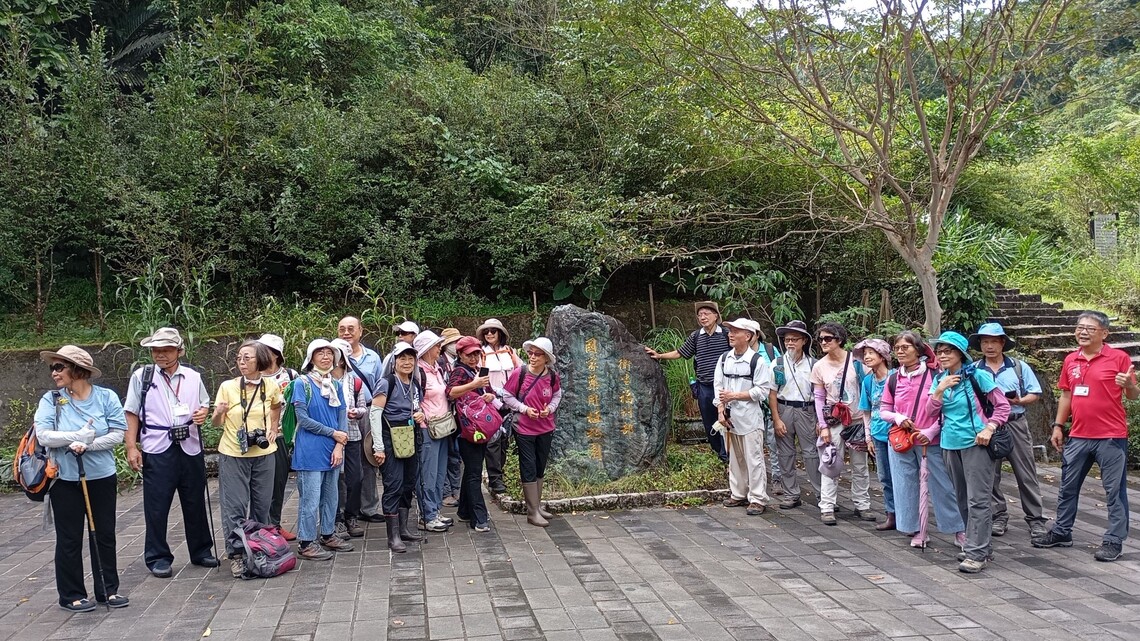 草根共學團員們在石碑前合影，氣氛愉快，背景為茂密綠樹與自然景觀。圖片