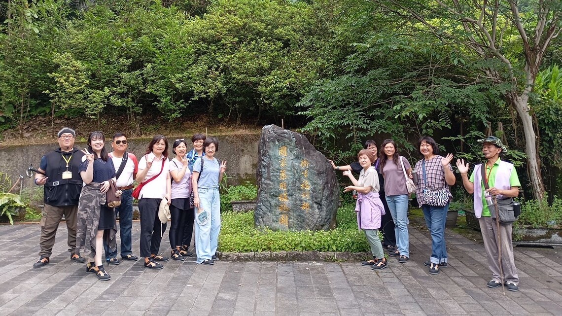平溪衛生所約 10 位成員於「衛生福利部 國家藥用植物園」石碑前合影。團員身著輕便服裝與便鞋，部分人手指向石碑，其他人則比出 V 手勢或揮手致意。石碑立於綠色灌木與樹木圍繞之間，地面為灰磚步道，整體畫面明亮清新，展現健康、活力與團隊間的融洽氛圍。
