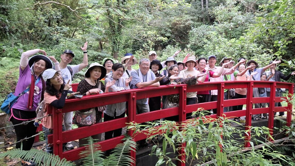萬華社大青草班 20 位成員站在森林中的紅色木橋上合影，笑容燦爛，背景為濃密林木與綠意。
