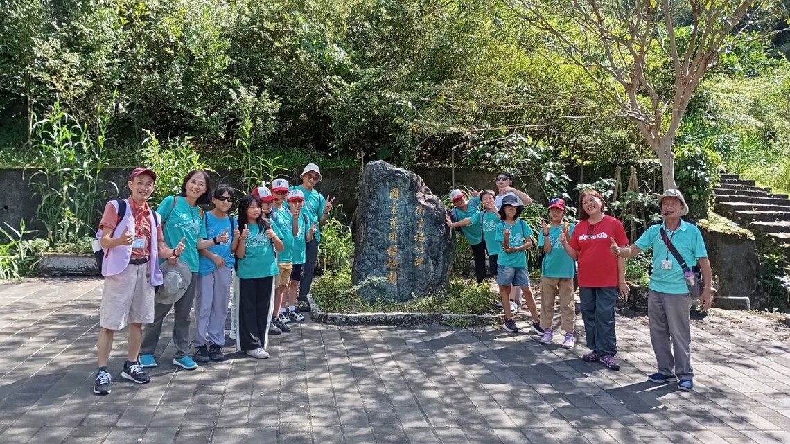 平溪國小15人參訪國家藥用植物園，學生與老師在石碑前合影，背景為綠樹環繞。圖片