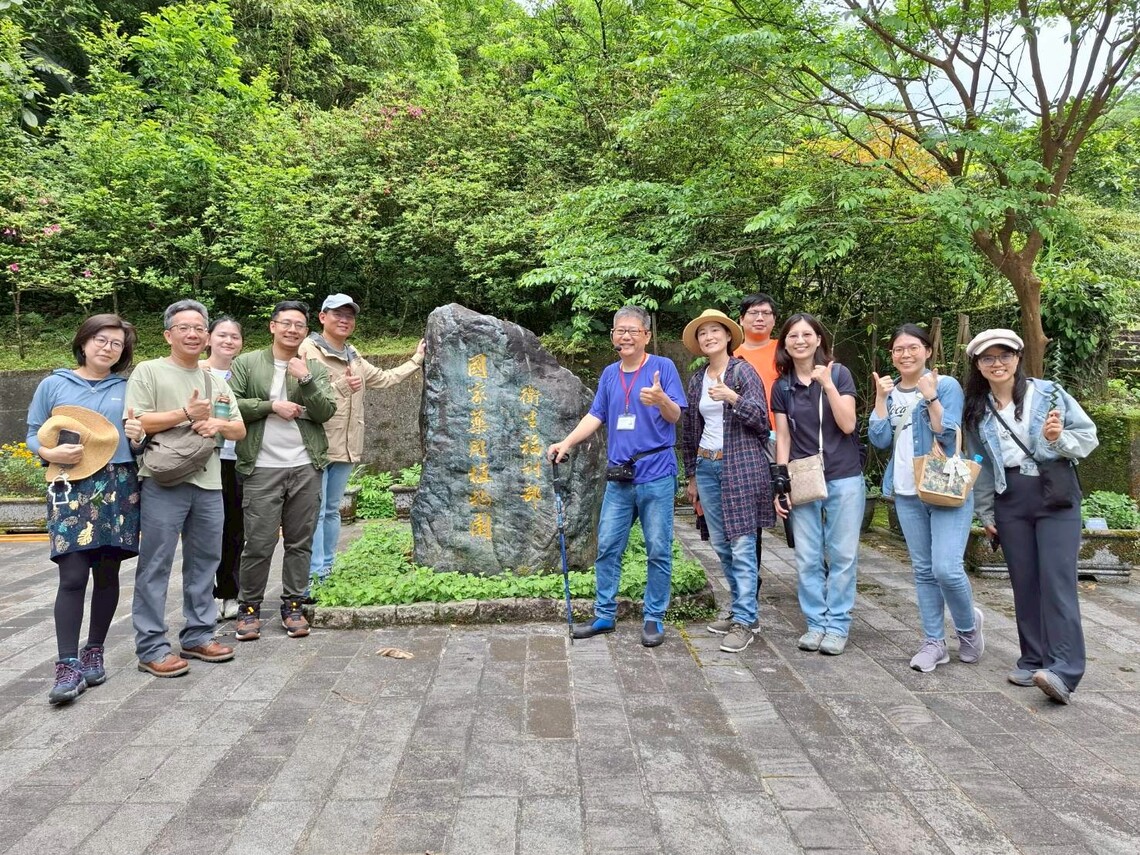 參訪者們站在刻有「衛生福利部國家藥用植物園」文字的大石頭旁邊，一隻手豎起大拇指