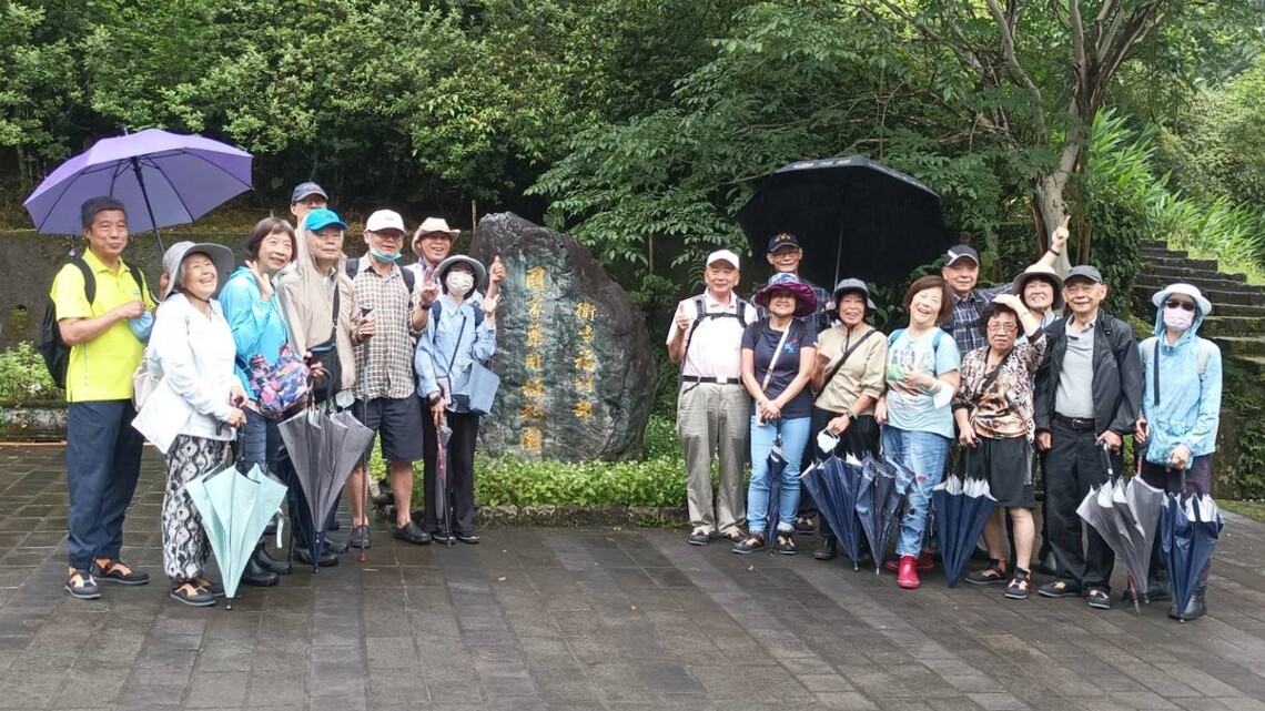 文山社區大學科學養生班共 18 位成員參訪「衛生福利部 國家藥用植物園」，於園區標誌性石碑前合影。團員年齡層偏中高齡，穿著輕便衣物與遮陽帽或雨具，部分人手持或撐開雨傘，站立於濕潤的石板地面上。石碑兩側為茂密樹林與階梯，整體氛圍溫暖友善，展現對健康與自然的關注與參與。