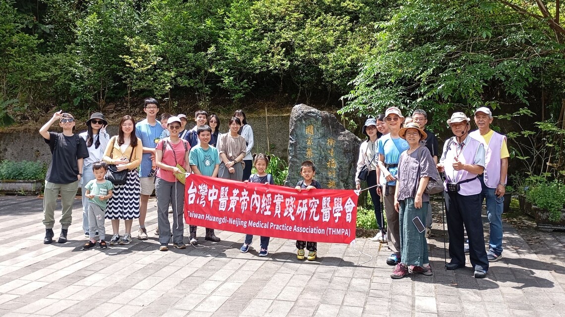 照片中約二十五人，包括成人與多位孩童，於戶外合影。眾人站在「衛生福利部 國家藥用植物園」石碑前，部分成員手持紅色布條，寫有「台灣中醫黃帝內經實踐研究醫學會 Taiwan Huangdi-Neijing Medical Practice Association (THMPA)」字樣。前排有三位兒童站在布條後方，神情活潑。成員服裝多樣，包含休閒裝與遮陽帽，背景為濃密綠樹與自然步道，整體呈現融合家庭與中醫文化交流的輕鬆參訪氛圍。