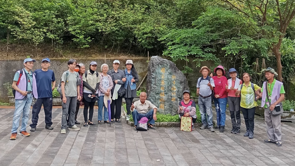 照片中共有十五人，為平溪老人會成員，在 2025 年 5 月 1 日進行參訪活動時於戶外合影。大部分人穿著便服、帽子並攜帶隨身物品，站立或蹲坐於一塊題有「衛生福利部 國家藥用植物園」字樣的大石碑前，背景是濃密的綠色植栽與樹木。地面為灰色磚石鋪面，陽光和煦，現場氛圍愉悅溫馨。圖片