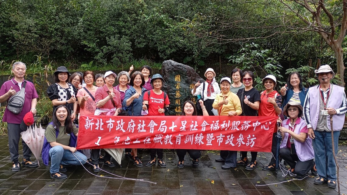 30位志工於國家藥用植物園石碑前合影，舉著紅色橫幅，現場氣氛熱絡愉快。圖片