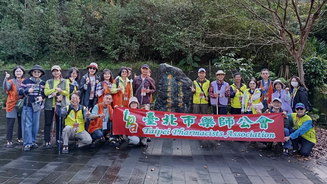 臺北市藥師公會20人參訪國家藥用植物園，成員舉著「臺北市藥師公會」橫幅於石碑前合影，氣氛熱烈愉快。圖片