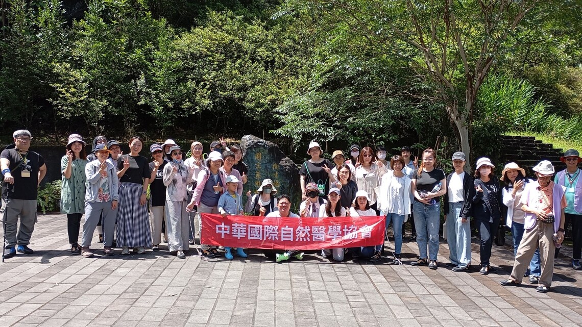 30多人於藥用植物園石碑前合影，前排舉紅色橫幅，背景為林木與階梯，氣氛熱絡歡樂。