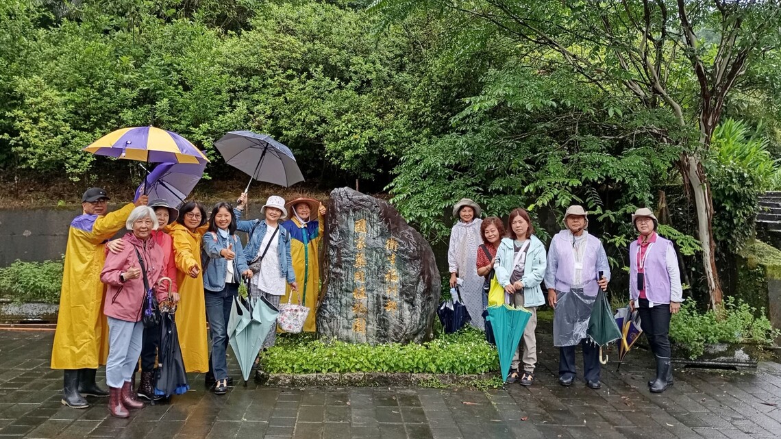 民眾團體共 10 位成員參訪「衛生福利部 國家藥用植物園」，於園區石碑前合影。當日為雨天，成員中有多人穿著黃色與紫色雨衣，部分人撐傘或手持摺疊傘，也有人穿著防水靴。眾人站立於濕潤的磚石步道上，背景為繁茂的樹木與濃綠植栽，畫面溫馨自然，呈現即使在雨中也不減熱情的參訪情景。